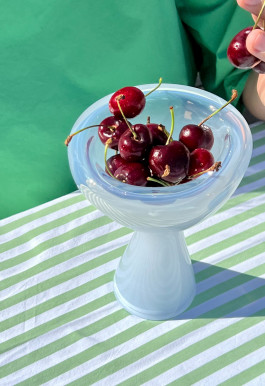 A light blue glass vase standing on a table. The vase is filled with cherries. Logo Progressive Present