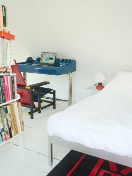 Minimalist bedroom with a white bed, modern blue desk, red chair, and a bookshelf filled with design books – contemporary interior inspiration. Logo Progressive Present