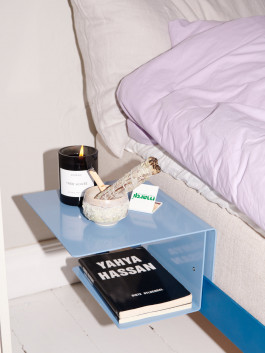 Modern floating bedside table in blue with a book, scented candle, and wellness accessories next to a bed with lilac bedding – minimalist bedroom decor. Logo Progressive Present