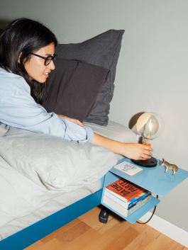 Woman in bed reaching for a modern bedside lamp on a blue floating nightstand with books and decorative items – cozy and minimalist bedroom design. Logo Progressive Present