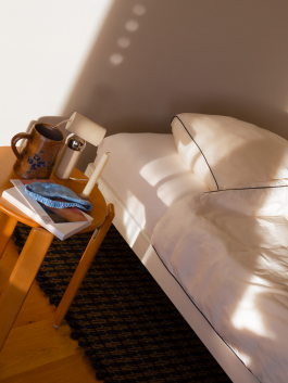 Cozy bedroom with warm sunlight, a wooden bedside table, books, and a ceramic mug next to a bed with white bedding – inviting minimalist interior. Logo Progressive Present