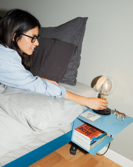 Woman in bed reaching for a modern bedside lamp on a blue floating nightstand with books and decorative items – cozy and minimalist bedroom design. Logo Progressive Present