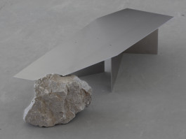 Sculptural table with angled brushed metal top resting on a natural stone base, minimalist industrial design Logo Progressive Present
