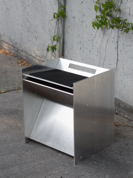 Stainless steel stool or side table with angular construction and black seat, positioned on concrete ground against a textured wall with climbing plants Logo Progressive Present