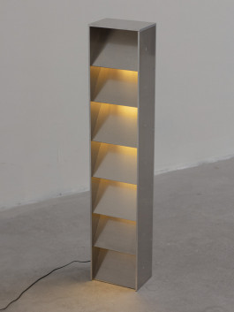 Tall brushed metal floor lamp with stepped interior shelves illuminated in warm light Logo Progressive Present
