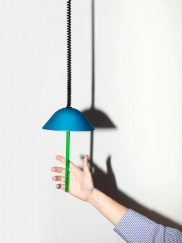 Blue pendant lamp with green detail and a hand reaching towards it, casting a strong shadow on a white wall Logo Progressive Present