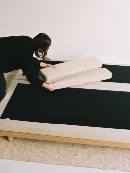 A woman adjusting foldable cushions on a wooden daybed with a black fabric base. Logo Progressive Present