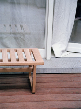 Detail view of a wooden slatted bench on a red timber deck in front of a glass door. Logo Progressive Present