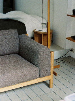 Detail view of a modern armchair in grey upholstery with a wooden frame, next to a bed and wooden floor lamp. Logo Progressive Present