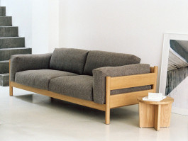 Minimal wooden sofa with grey cushions in a calm, light-filled interior next to a small round side table. Logo Progressive Present