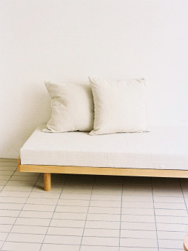 Minimal wooden daybed with two light-colored cushions in front of a white wall on tiled flooring. Logo Progressive Present