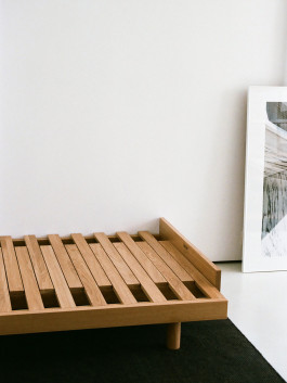 Minimalist solid wood bed frame with wide slats on a dark rug. Logo Progressive Present