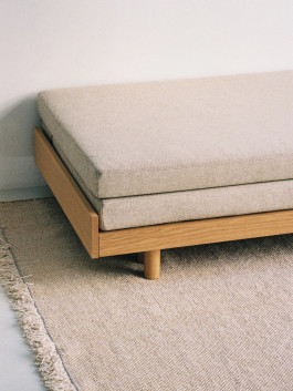 Detail view of a minimalist wooden daybed with beige cushions on a woven rug. Logo Progressive Present