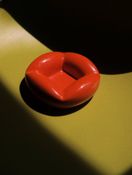 Glossy red ceramic ashtray in sculptural form, placed on an olive green surface with dramatic lighting. Logo Progressive Present