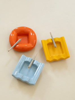 Three colorful ceramic ashtrays—red, yellow, and light blue—each holding a rolled joint, arranged on a pale surface. Logo Progressive Present