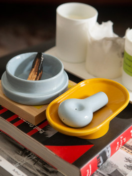 Light blue ceramic pipe and matching incense holder on a yellow tray, placed on a stack of books next to candles. Logo Progressive Present