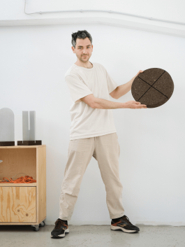 Designer holding a round cork tabletop with cross-cut detail, standing in a minimal studio. Logo Progressive Present