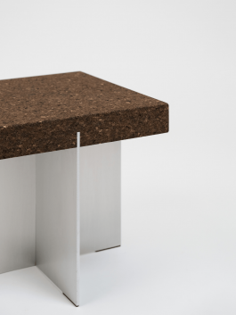 Close-up of a square stool with a thick cork top and brushed aluminum base against a white background. Logo Progressive Present