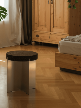 Minimalist stool with dark round cork seat and cross-shaped aluminum base in a wooden-floored bedroom. Logo Progressive Present