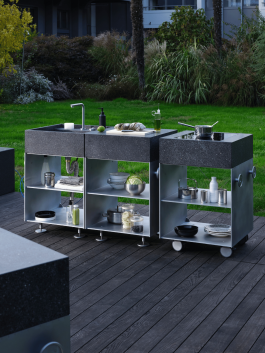 Outdoor kitchen modules with dark stone countertops and open stainless steel shelving, set on a wooden deck. Logo Progressive Present