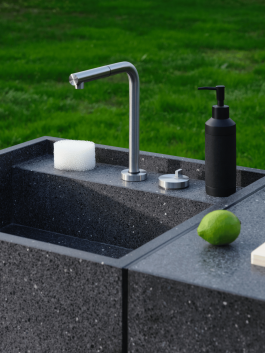 Close-up of a stainless steel faucet and black soap dispenser on a dark stone outdoor kitchen sink. Logo Progressive Present