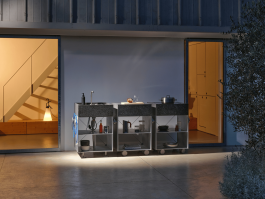 Three modular outdoor kitchen units with dark stone countertops in front of an open building at dusk. Logo Progressive Present